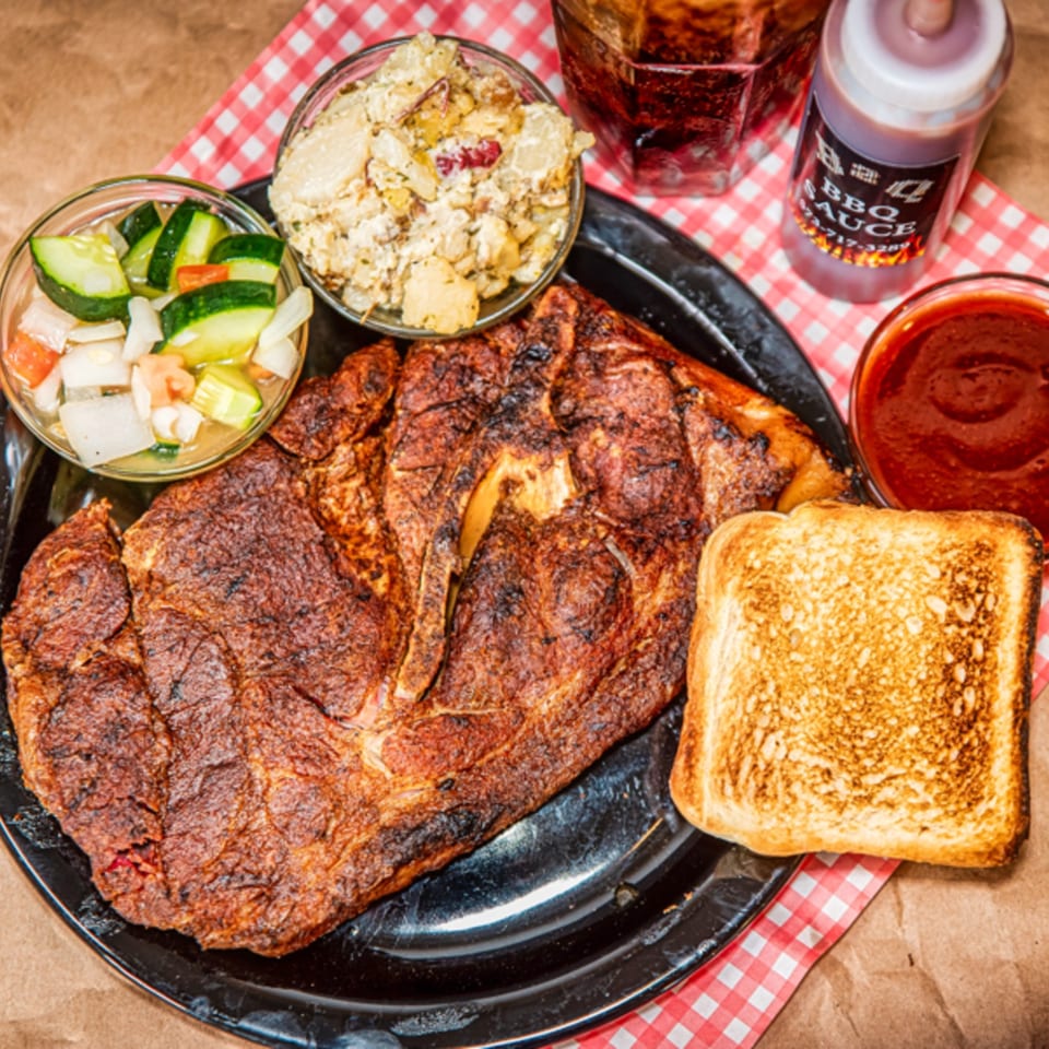 Grilled meat with sides of potato salad, cucumber salad, toast, and BBQ sauce.