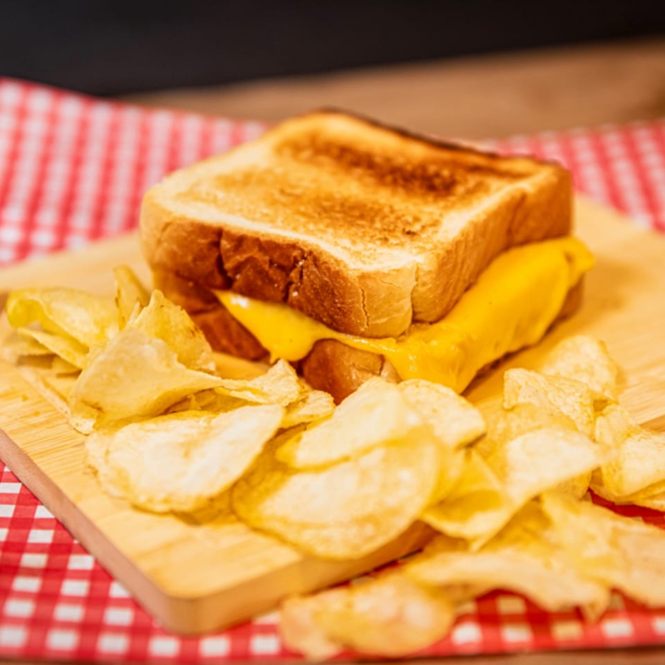 Grilled cheese sandwich with melted cheese and potato chips on a wooden board with a red and white checkered cloth.