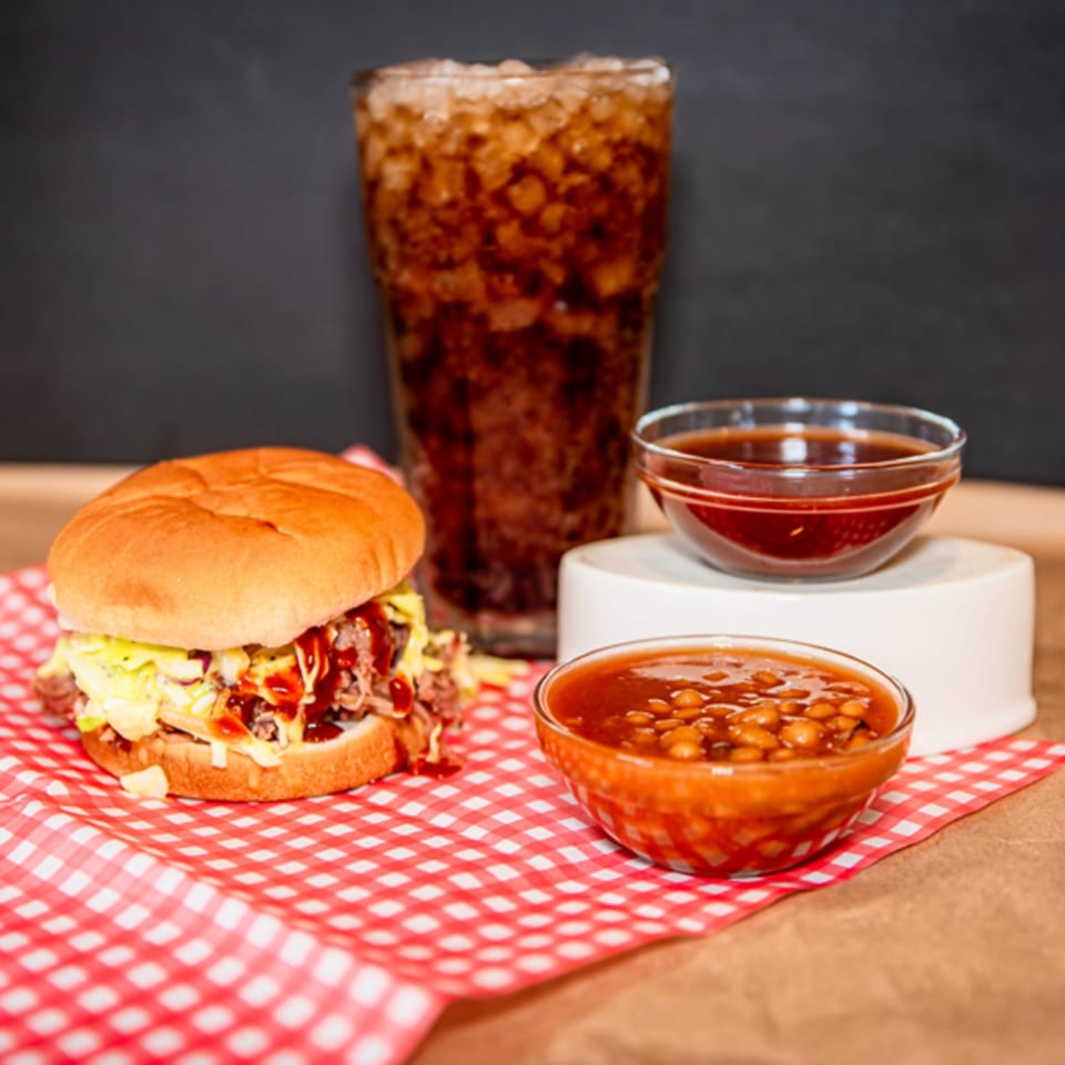 A pulled pork sandwich, baked beans, barbecue sauce, and a soda on a red checkered napkin.