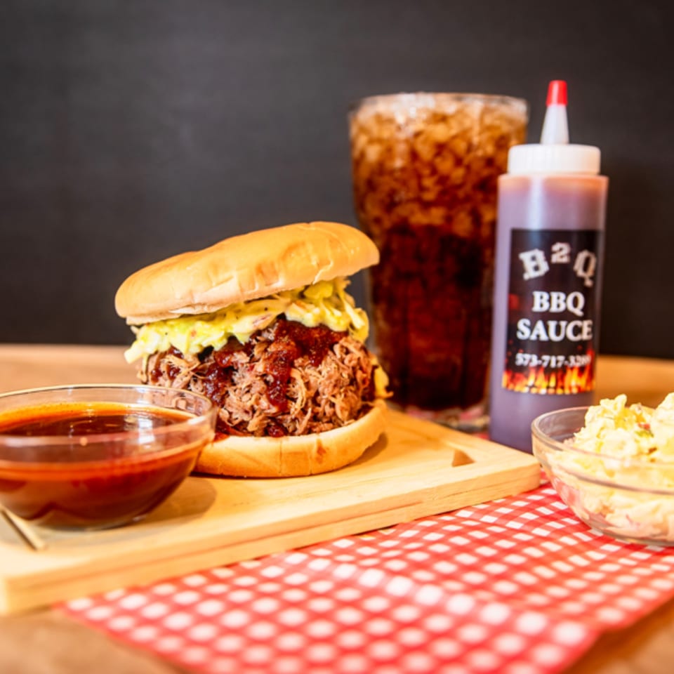 Pulled pork sandwich with coleslaw, BBQ sauce, and a drink on a checkered tablecloth.
